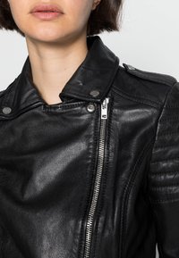 Černá kožená motocyklová bunda se zapínáním na zip, patentními knoflíky a texturovanými detaily na ramenou. Hladký povrch s přiléhavým střihem.