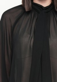Donna che indossa una blusa nera trasparente con un dettaglio legato al collo e capelli castani lisci che le ricadono sulle spalle.