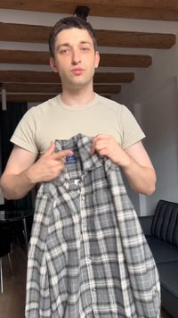 Νεαρός άνδρας σε εσωτερικό χώρο, κρατώντας και δείχνοντας ένα γκρι καρό πουκάμισο με κουμπιά, φοράει μπεζ μπλουζάκι, με ξύλινες δοκούς στην οροφή.