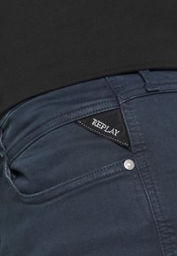 Mörkblå denimjeans med en texturerad yta, med en ficktagg som har "REPLAY"-logotyp och en silverfärgad knappdetalj.