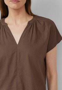 Haut court marron à manches courtes avec un décolleté en V, présentant des détails froncés au niveau du col et une coupe décontractée. Tissu lisse et texturé.