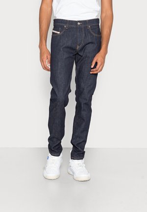 Jeans de mezclilla azul oscuro con corte ajustado, con costuras naranjas, cierre de botón en la parte frontal y cinco bolsillos, combinados con zapatillas blancas.