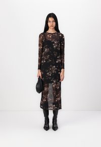 Robe noire transparente avec un motif floral dans des tons bruns, à manches longues et fente haute. Le mannequin tient un sac noir et porte des bottes noires à talons pointus.