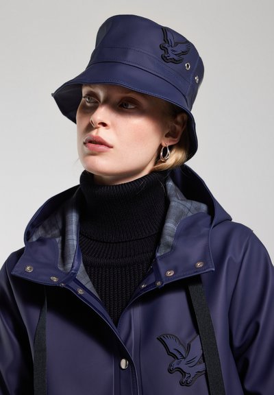 Marineblaue wasserfeste Jacke mit Kapuze, Frontlogo, Druckknöpfen und gefüttertem Kragen. Kombiniert mit einem schwarzen gerippten Rollkragenpullover.
