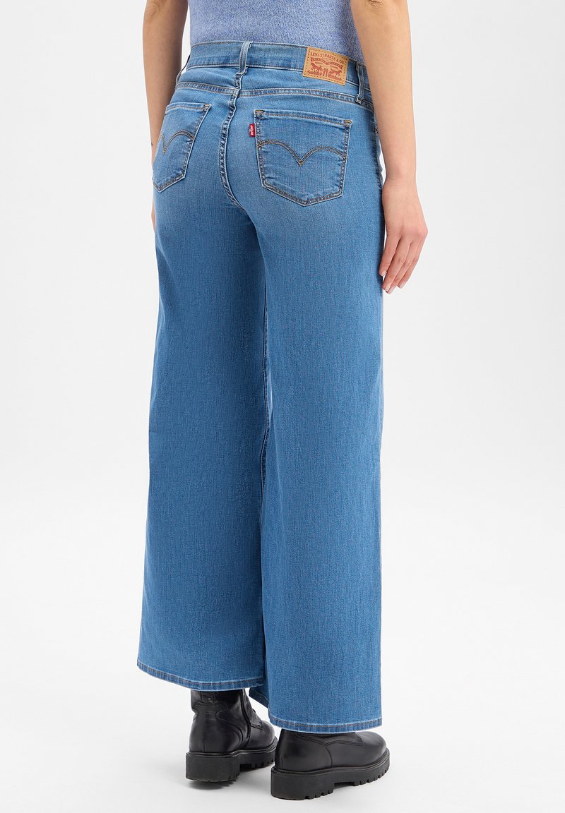 Person wearing high-waisted, wide-leg blue jeans with back pockets and black chunky boots, standing on a white background.