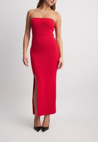 Rotes, geripptes, trägerloses Maxikleid mit einem Seitenschlitz, das eine enganliegende Silhouette und glatte Textur aufweist, kombiniert mit schwarzen spitzen High Heels.