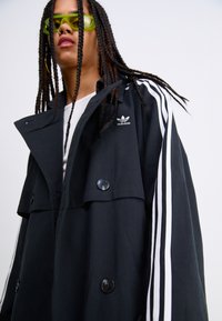 Manteau trench noir oversize avec des rayures blanches le long des manches, doté d'une fermeture à double bouton et d'un logo Adidas brodé sur la poitrine.