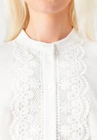 Blusa bianca con un pannello in pizzo con motivi floreali al centro, decorata con piccoli bottoni e un colletto tondo.