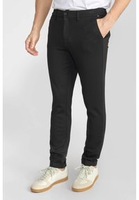 Pantalons noirs ajustés en tissu lisse. Dotés de poches avant et d'une fermeture à bouton. Portés avec des baskets blanches.