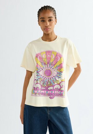 Junge Frau mit geflochtenem Haar, die ein cremefarbenes T-Shirt mit einer bunten Sonne- und Mond-Grafik und dem Schriftzug „Wrangler“ trägt, kombiniert mit blauen Jeans.