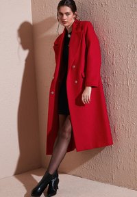 Red wool coat with a tailored design, double-breasted front, and silver buttons. Smooth texture and long length, paired with a black dress.