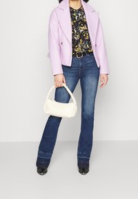 Femme portant une veste lavande, un chemisier fleuri, un jean bleu et des chaussures noires, tenant un sac à main blanc tressé sur un fond uni.