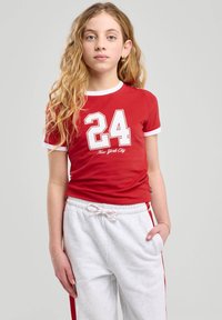 Camiseta de algodón roja con ribete blanco, que presenta un gran "24" blanco y el texto "New York City". Combinada con pantalones de chándal grises con rayas laterales rojas.