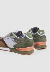 Sneakers verdi e marroni con una combinazione di materiali in suede e mesh, caratterizzati da accenti bianchi e un dettaglio in rame sul tallone. Anno del modello "1973" esposto.