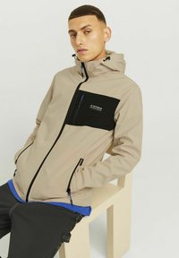 Beige hooded jacket with a black chest pocket, contrasting zipper details, and a smooth texture. Worn over a blue shirt.