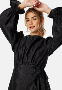 Robe noire avec de longues manches ballon, faite en tissu froissé. Ses caractéristiques notables comprennent une taille cintrée et une jupe ample et fluide.
