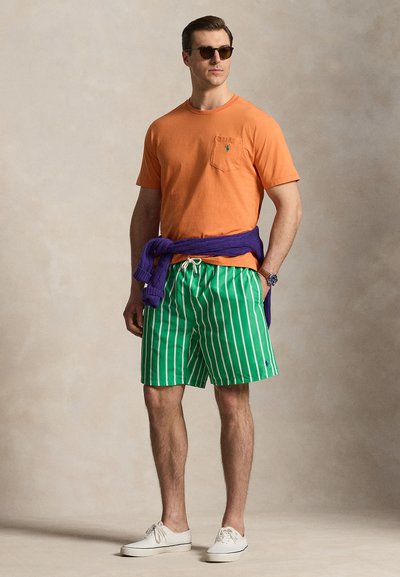 Vyras, dėvintis oranžinį marškinėlį, žaliai baltomis juostelėmis dryžuotas trumpikes, baltus sportinius batelius, dėvintis akinius nuo saulės ir violetinį megztinį, surištą aplink juosmenį, stovintis rankomis kišenėse.