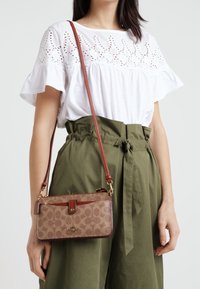 Brązowa torba na ramię z wzorem, z czerwoną skórzaną rączką, o prostokątnym kształcie i zapięciem na zatrzask. Torba ma subtelny detal z logo.