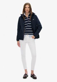 Jeune femme portant une veste bleu marine, un haut rayé bleu marine et blanc, un pantalon blanc et des mocassins marron foncé, debout devant un fond blanc uni.