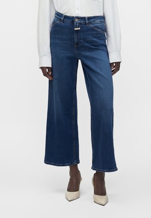 Personne portant un jean bleu taille haute à jambes larges, une chemise blanche à manches longues rentrée, et des talons beiges à bouts pointus sur un fond uni.