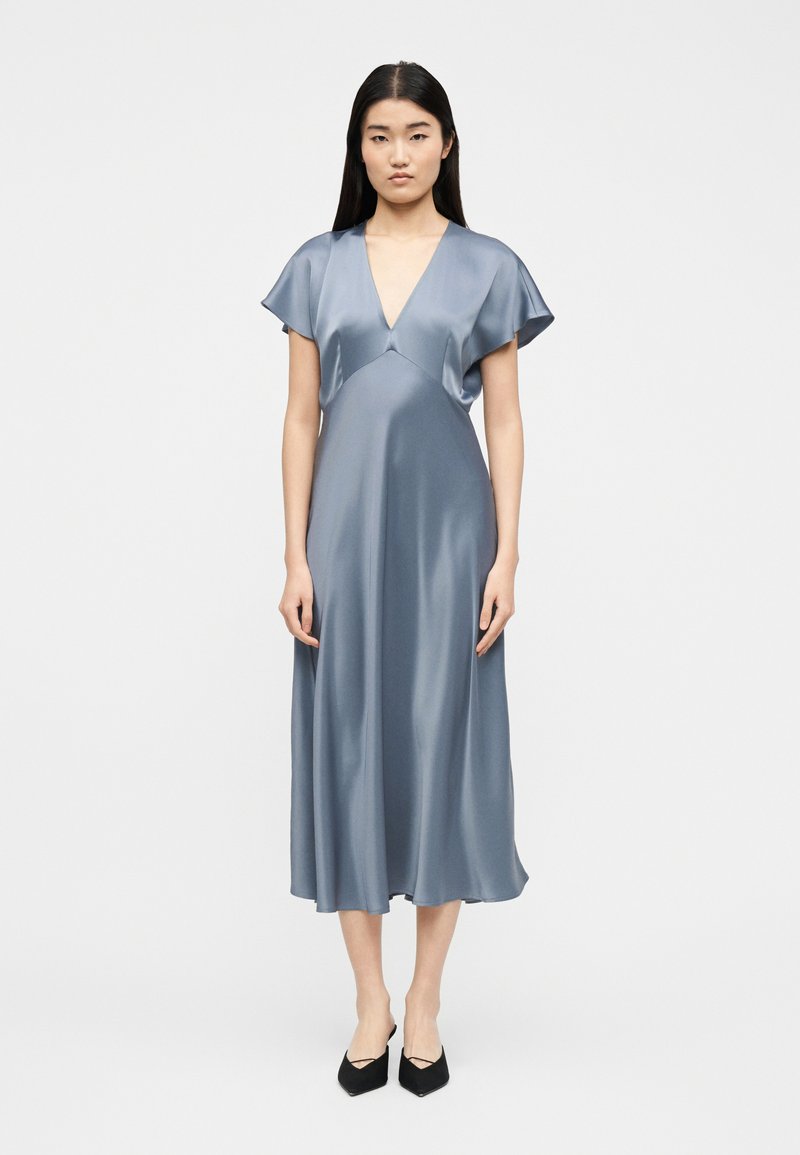 Νεαρή γυναίκα  που φοράει ένα λαμπερό μπλε-γκρι midi φόρεμα με V-λαιμόκοψη, κοντά φουσκωτά μανίκια και μαύρα μυτερά παπούτσια.