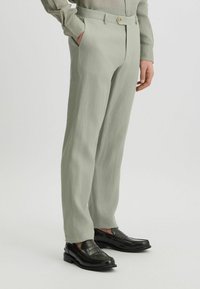 Hellgrüne Hose aus einem strukturierten Stoff, mit geradem Schnitt und Knopfverschluss. Kombiniert mit schwarzen Slippern.
