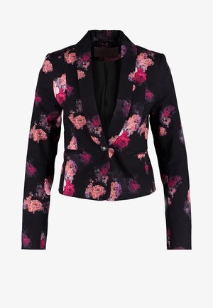 Vrouw draagt beige leren broek, zwarte blazer met bloemenmotief, zwart topje, zwarte enkellaarzen en houdt een bordeauxrode clutch vast.