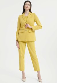Completo giallo su misura con giacca a bottone singolo, due tasche frontali e pantaloni a sigaretta abbinati. Scarpe nude con tacco alto completano l'outfit.