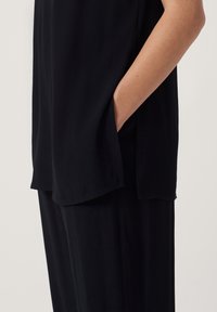 Svart avslappnad topp med sidoflik och ficka, tillverkad av ett mjukt tyg med slät textur. Framhävd av en diskret design.