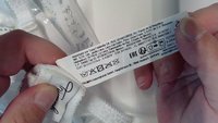 Biely textil s čipkovaným lemovaním, prezentujúci štítok so starostlivosťou. Štítok obsahuje pokyny na pranie a podrobnosti o výrobcovi, vrátane ikon a textu.