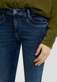 Jeans de mezclilla azul oscuro de corte slim, con bolsillos delanteros, un cierre de botón visible y una textura suave, combinados con una blusa verde oliva.