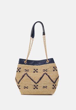 Sac à main beige tissé avec motifs géométriques foncés, garniture en cuir bleu marine et poignées en chaîne dorée avec coussinets d'épaule bleu marine sur fond blanc.