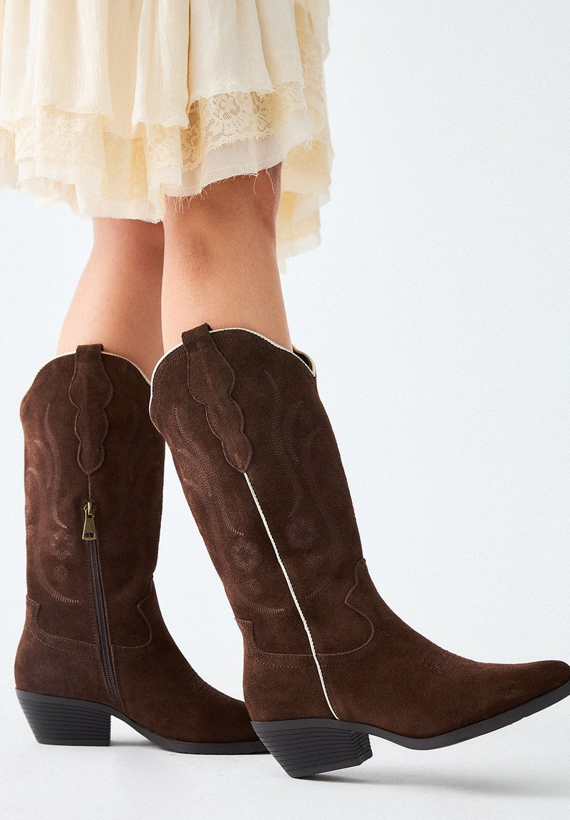 Botas de vaquero de ante marrón con punta afilada, costuras decorativas, cremallera lateral y detalles en blanco a lo largo del borde superior. Diseño de tacón bajo.
