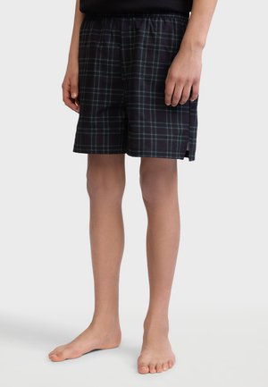 Person trägt schwarze und graue karierte Shorts mit einem elastischen Bund und steht barfuß vor einem schlichten Hintergrund.