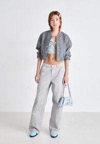 Cazadora bomber corta gris con botones, top corto azul claro, pantalones anchos grises y zapatos de tacón azul claro, sosteniendo un bolso estampado.