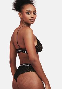 Set di lingerie in pizzo nero composto da un bralette con spalline sottili e un thong abbinato, accentuato da un band con logo. Texture liscia e design delicato.
