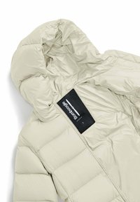 Off-white pufferjack met een capuchon, gekwilt textuur en een zichtbaar zwart etiket met witte tekst binnenin de kraag. Kraagzipper zichtbaar.