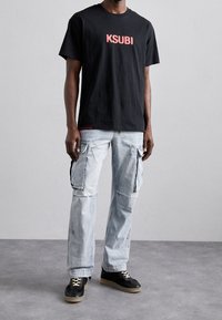 Černé bavlněné tričko s logem "KSUBI" v červené barvě. Světle modré cargo kalhoty s předními kapsami. Černé tenisky s béžovými podešvami.