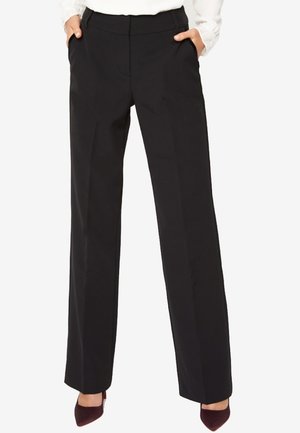 Frau trägt schwarze, gerade geschnittene Hosen mit den Händen in den Taschen und burgunderrote spitze Schuhe, weißes Oberteil teilweise sichtbar.