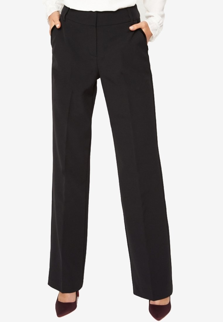 Femme portant un pantalon noir à jambes droites avec les mains dans les poches et des escarpins bordeaux à bout pointu, haut blanc partiellement visible.