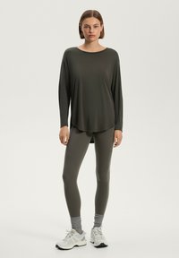Maglietta a maniche lunghe di colore verde scuro con scollo rotondo, abbinata a leggings aderenti verdi scuri. Il modello indossa sneakers bianche con dettagli grigi.