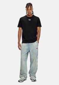 T-shirt en coton noir avec un logo circulaire sur la poitrine, associée à un jean en denim oversize bleu clair et des baskets argentées.