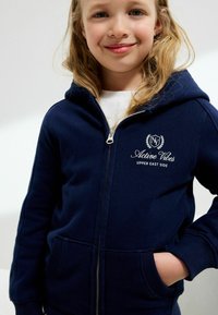 Barn der smiler, iført en navy blå zip-up hættetrøje med teksten og logoet "Active Vibes Upper East Side", med hænderne i lommerne foran en ensfarvet baggrund.