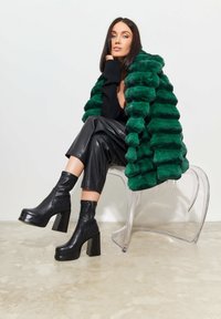 Groene nep-bontjas met horizontale strepen, zwarte gebreide top, leren broek en stevige platte enkelboots, zittend op een doorzichtige stoel.