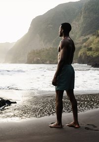 Une personne torse nu se tient sur une plage de sable, portant un maillot de bain teal, faisant face aux vagues de l'océan et à la côte rocheuse, sous une lumière douce et naturelle.