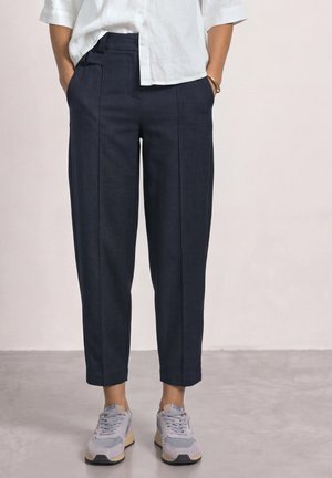 Personne portant un pantalon bleu marine sur mesure jusqu’aux chevilles, une chemise blanche boutonnée et des baskets grises, debout sur un sol gris avec les mains dans les poches.
