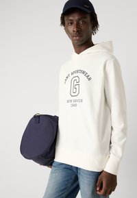 Sudadera blanca con capucha y gráfico azul marino que dice "GANT SPORTSWEAR NEW HAVEN 1949." El modelo sostiene un bolso circular azul marino y lleva puesto unos jeans azules.