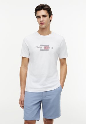 Νεαρός άνδρας που φοράει λευκό T-shirt Tommy Hilfiger με λογότυπο και ανοιχτό μπλε σορτς, στέκεται μπροστά σε απλό λευκό φόντο.