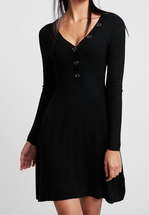 Kobieta w czarnej, prążkowanej, dzianinowej sukience z długimi rękawami, dekoltem w szpic i czterema guzikami z przodu.