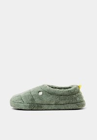 Chausson d’intérieur doux et vert, doublé en polaire, avec un motif matelassé, un petit talon et une petite languette jaune à l’arrière.